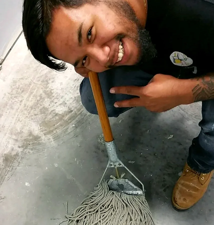 A Faithful Cleaning Service crew member kneeling on a concrete floor on a job site, smiling next to a string mop