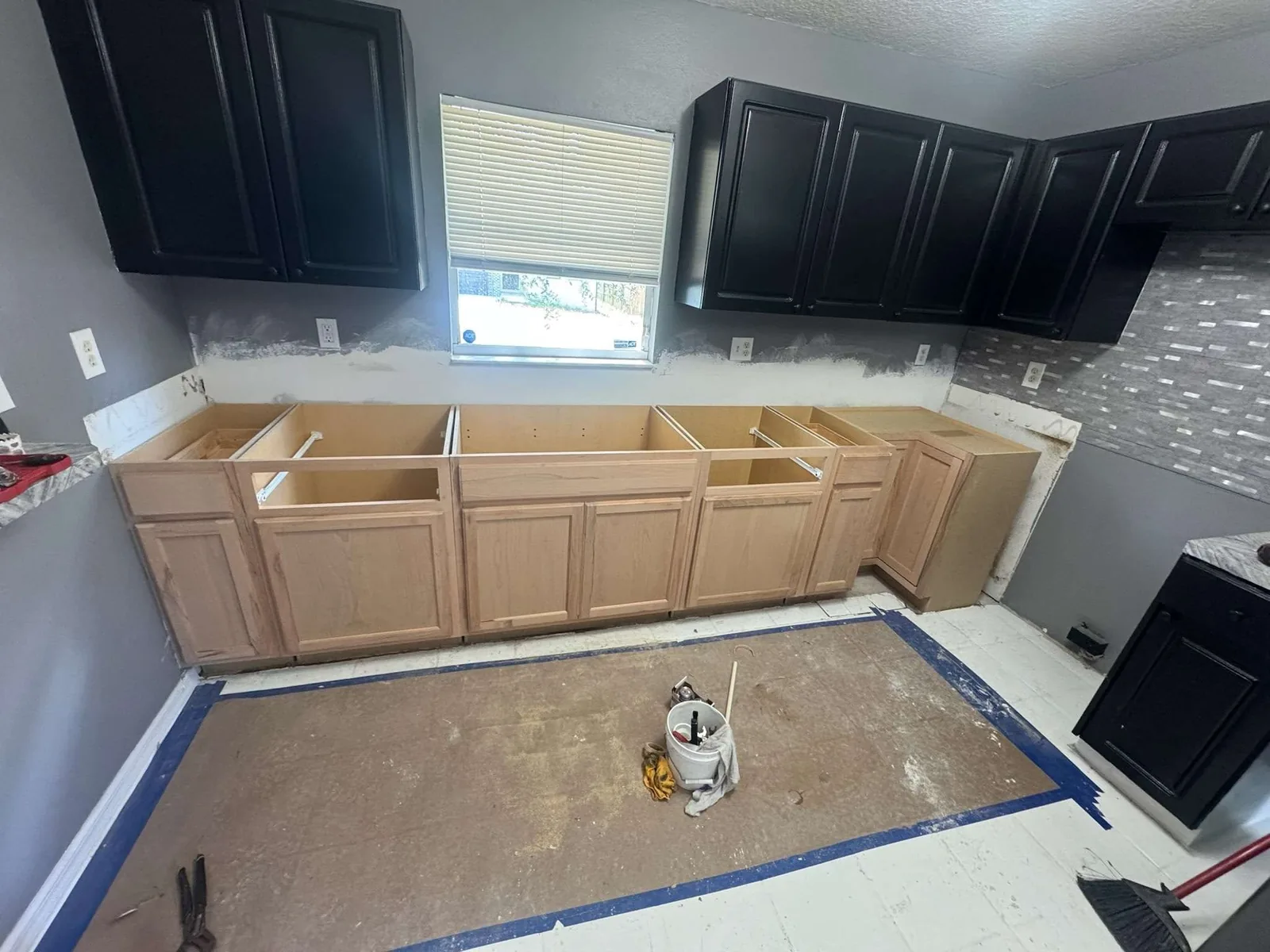 A wide view of the kitchen mid-rehab with black upper cabinets, raw base cabinets and floor prep underway